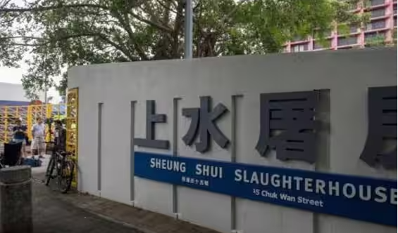 Foto del mattatoio di Sheung Shui (Hong Kong): un muro bianco con alcuni caratteri cinesi; sotto ai caratteri una banda blu con il nome del mattatoio in bianco.