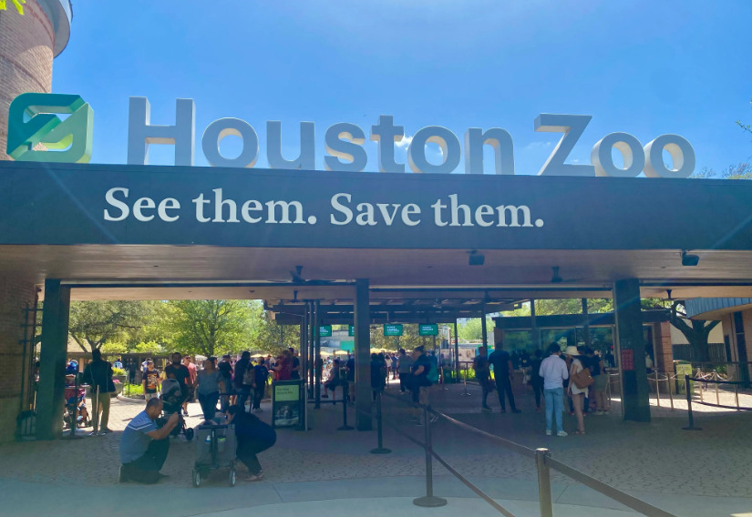 Foto dell'ingresso dello zoo di Houston. Nella parte inferiore, diversi visitatori stanno entrando o attendendo di entrare. Nella parte superiore, una grossa insegna con la scritta "Houston Zoo. See them. Save them".